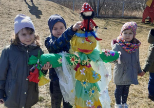 Troje dzieci trzyma kukłę Pani Wiosny, chłopiec posadził maskotkę Krasnala Hałabały na jej głowie.